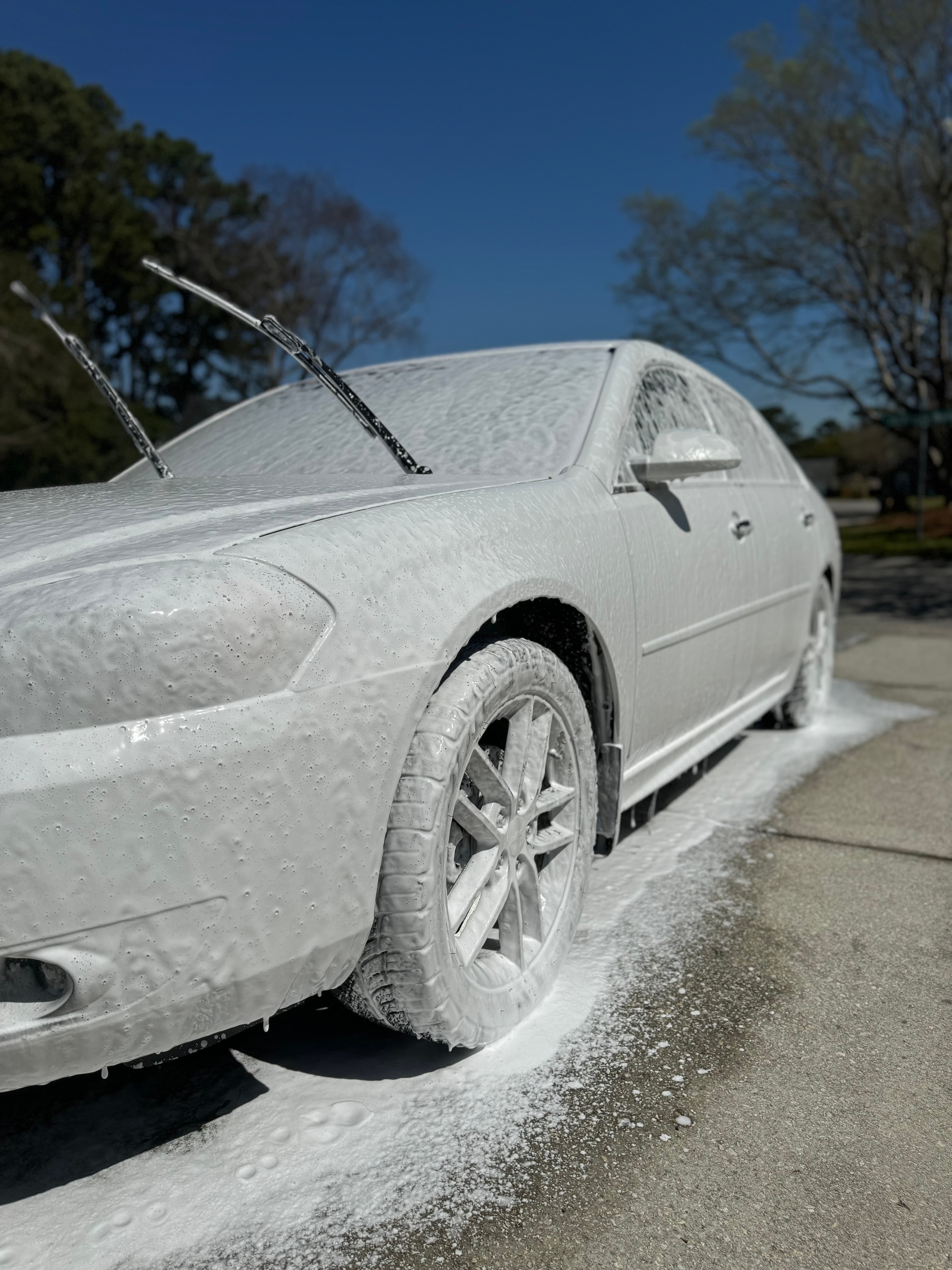 Silver sedan covered in thick snow foam during a foam-bath wash under blue sky